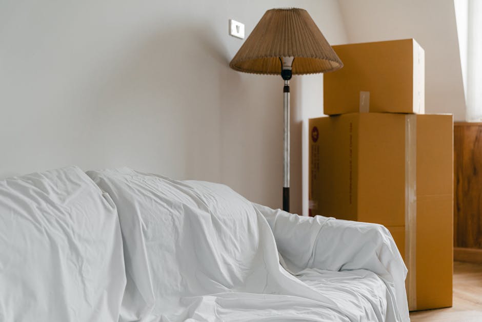 Interior view of a room showing a white sofa with a wrinkled fabric cover positioned against a plain light-colored wall. To the right of the sofa, three stacked cardboard boxes of varying sizes are pl