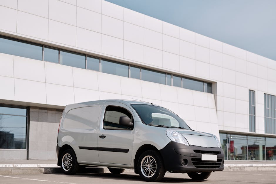 A small silver van is parked on the street in front of a modern white building with large windows. The van is positioned parallel to the building, with its driver’s side facing the camera. The van's side door is closed, and the vehicle appears to be used for home relocation or furniture transport. The surrounding environment is well-lit under an overcast sky, with the pavement clear and free of obstructions. The building behind features sleek, contemporary architecture with horizontal and vertical window panels. This image is intended to support content about house removals and parking tips, as provided by Man With a Van Balham, showcasing a typical vehicle used in packing, loading, and transporting household items during a moving process.
