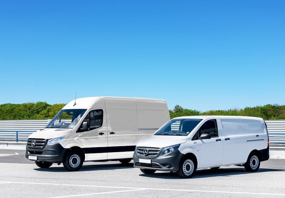 Two Mercedes-Benz commercial vans, a larger high-roof panel van and a smaller standard panel van, are parked side by side on a paved roadway near a metal barrier with a green treeline and clear blue sky in the background. The larger van, positioned to the left, is white with black accents on the grille and bumper, and features a sliding side door, rear doors, and a roof antenna, indicating its use for home relocation or furniture transport. The smaller van, to the right, is also white with similar black detailing, with its front windows rolled down slightly, suggesting recent loading or unloading activity. The scene is lit by bright daylight, with minimal shadows, and the empty road surface suggests a parking or staging area typical of moving logistics. This setting reflects professional moving services, such as those offered by Man With a Van Balham, highlighting the vehicles used for packing and transporting household items during residential relocations.