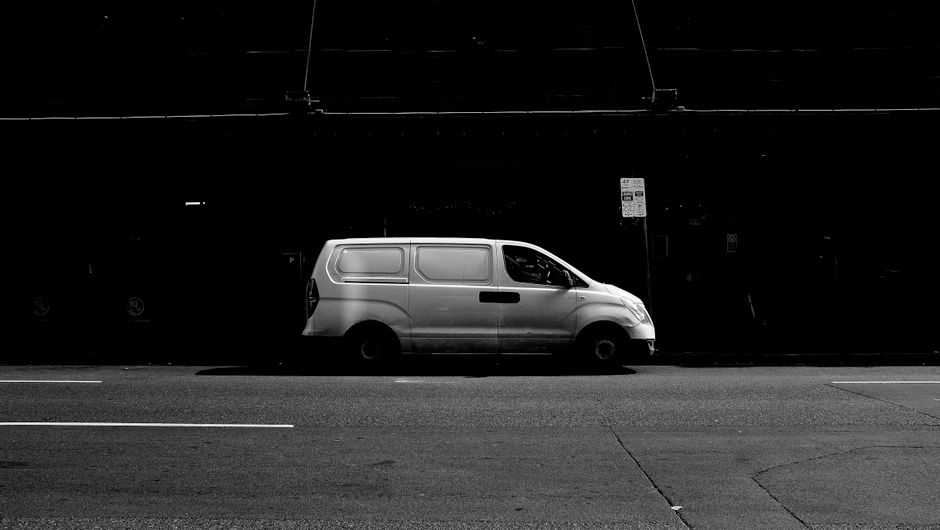 A small silver van is parked on the street in front of a modern white building with large windows. The van is positioned parallel to the building, with its driver’s side facing the camera. The van's side door is closed, and the vehicle appears to be used for home relocation or furniture transport. The surrounding environment is well-lit under an overcast sky, with the pavement clear and free of obstructions. The building behind features sleek, contemporary architecture with horizontal and vertical window panels. This image is intended to support content about house removals and parking tips, as provided by Man With a Van Balham, showcasing a typical vehicle used in packing, loading, and transporting household items during a moving process.