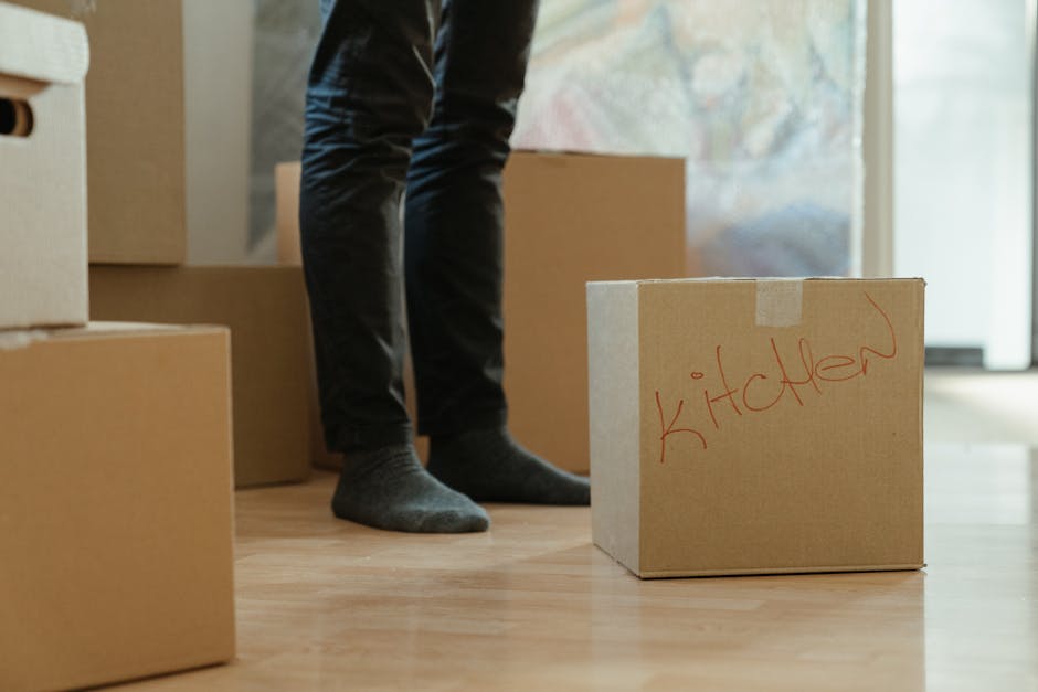 A person wearing dark pants and grey socks is standing inside a house near an open doorway, surrounded by several cardboard boxes used for house removals. One of the boxes, positioned in the foreground, has the word 'kitchen' written in red marker, indicating its contents related to the kitchen. The boxes vary in size, with some stacked and others placed on a wooden floor, ready for packing or transport as part of a home relocation process. The background shows a glimpse of the outside through the doorway, suggesting natural daylight illuminating the interior. The scene captures the packing and moving logistics involved in furniture transport and house removals, with visible packaging materials and a focus on organization and preparation for a smooth move, supported by [COMPANY_NAME] during the loading process.