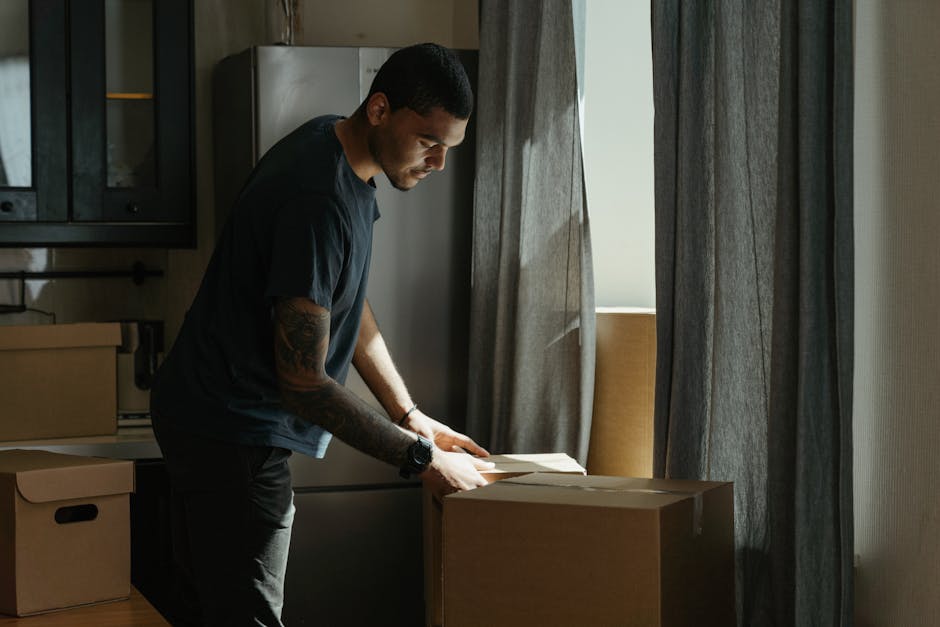 A man inspects or prepares packing materials on a cardboard box inside a room with natural light coming through partially open curtains. He is wearing a dark t-shirt and a watch, standing beside a kitchen counter with additional cardboard boxes and packaging supplies nearby. The room has a modern interior with cabinetry, a refrigerator, and neutral-toned curtains. This scene depicts a home relocation process, emphasizing packing and moving logistics as part of a house removal. The man with a van professional from Man With a Van Balham is visible managing packing tasks, contributing to efficient furniture transport and overall house move arrangement, supporting the detailed Balham station removals guide for flat moves.