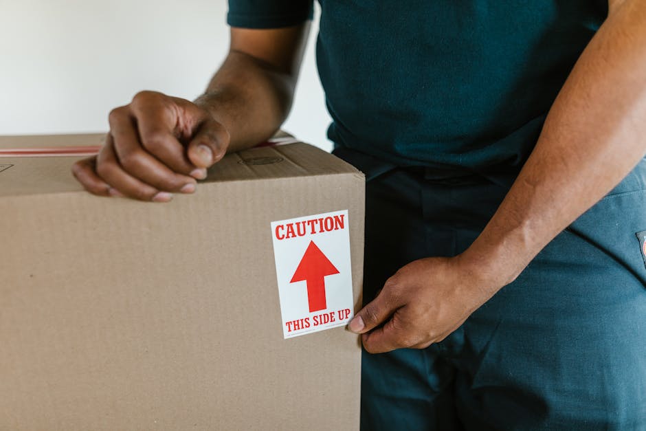A person wearing dark pants and grey socks is standing inside a house near an open doorway, surrounded by several cardboard boxes used for house removals. One of the boxes, positioned in the foreground, has the word 'kitchen' written in red marker, indicating its contents related to the kitchen. The boxes vary in size, with some stacked and others placed on a wooden floor, ready for packing or transport as part of a home relocation process. The background shows a glimpse of the outside through the doorway, suggesting natural daylight illuminating the interior. The scene captures the packing and moving logistics involved in furniture transport and house removals, with visible packaging materials and a focus on organization and preparation for a smooth move, supported by [COMPANY_NAME] during the loading process.