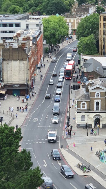 A view of a busy urban street near Tooting Bec Common, featuring a row of parked cars along the curb and several vehicles moving along the road, including a white van, a red double-decker bus, and various smaller cars. On the sidewalk, pedestrians are walking, some carrying bags, while others stand near the pavement edge. To the right, there is a historic-style building with a small tower and a white facade, adjacent to a residential property with a grey roof. Large green trees line the street, providing shade and adding greenery to the scene. In the background, multi-storey buildings and the canopy of Tooting Bec Common are visible, emphasizing the proximity to public parkland. During a typical home relocation or furniture transport operation, [COMPANY_NAME] would coordinate the loading process, with furniture and boxes carefully wrapped and arranged in the back of a van, ready for moving or delivery, supporting the logistical aspects of house removals in the Balham area.