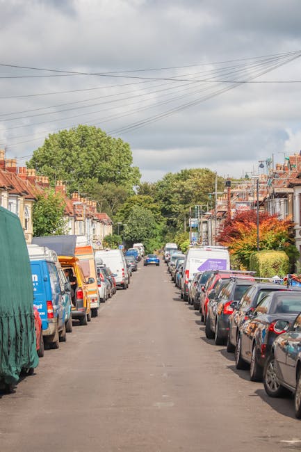 A residential street in Balham featuring parked cars lining both sides of the road, with a clear view down the street towards lush green trees and a partly cloudy sky overhead. On the left side, a van with a green protective cover is partially visible, parked near a house with a driveway. On the right side, various vehicles including small family cars and commercial vans are parked close to the pavement, some of which are part of a moving or removal process associated with house relocations. The scene captures a typical suburban neighbourhood setting, with overhead power lines running along the street and well-maintained houses with front gardens. The environment suggests a peaceful area often used for home removals or furniture transport, with the presence of a Man With a Van Balham service occasional in the context of logistical planning for house moves, highlighting the importance of careful vehicle access and parking during the loading process for efficient relocation.